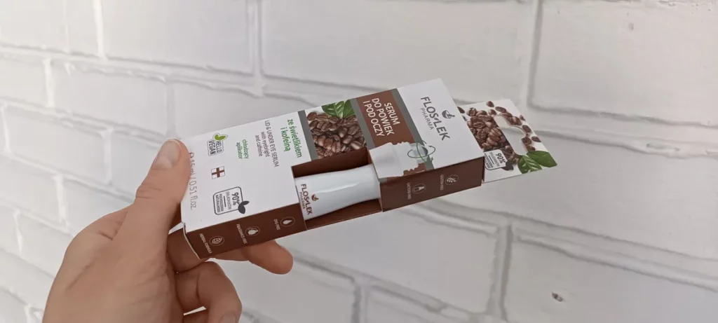Butelka serum w opakowaniu trzymana przez dłoń na tle białej, ceglanej ściany.