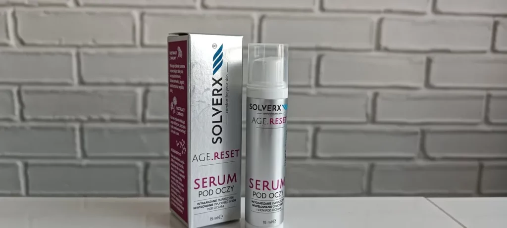 Butelka serum pod oczy stoi obok kartonika na białym blacie przed szarą ceglaną ścianą.