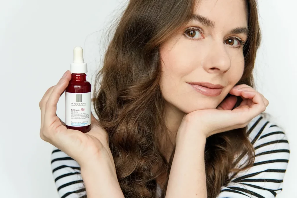 Kobieta trzyma butelkę serum z pipetą, prezentując ją z łagodnym uśmiechem na jasnym tle.