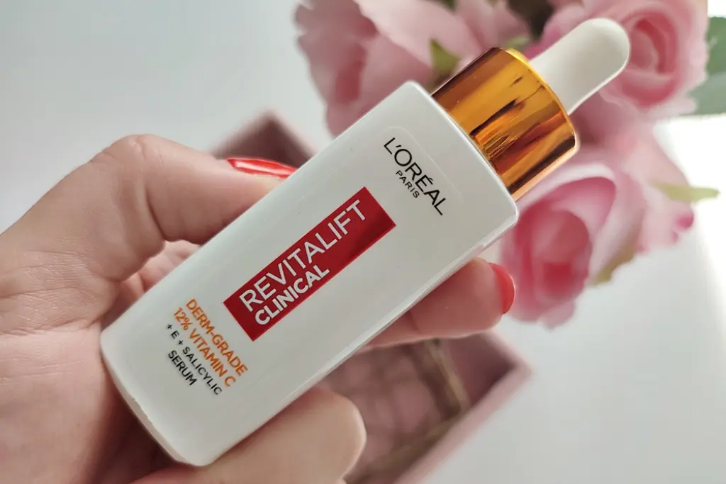 Butelka serum L'Oréal trzymana w dłoni, z kroplomierzem, na tle różowych róż.