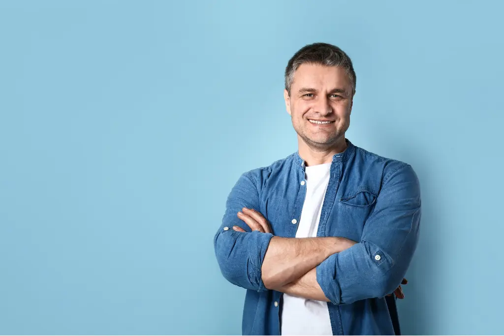 Mężczyzna stoi z założonymi rękami i uśmiecha się w luźnej koszuli jeansowej na jasnoniebieskim tle.