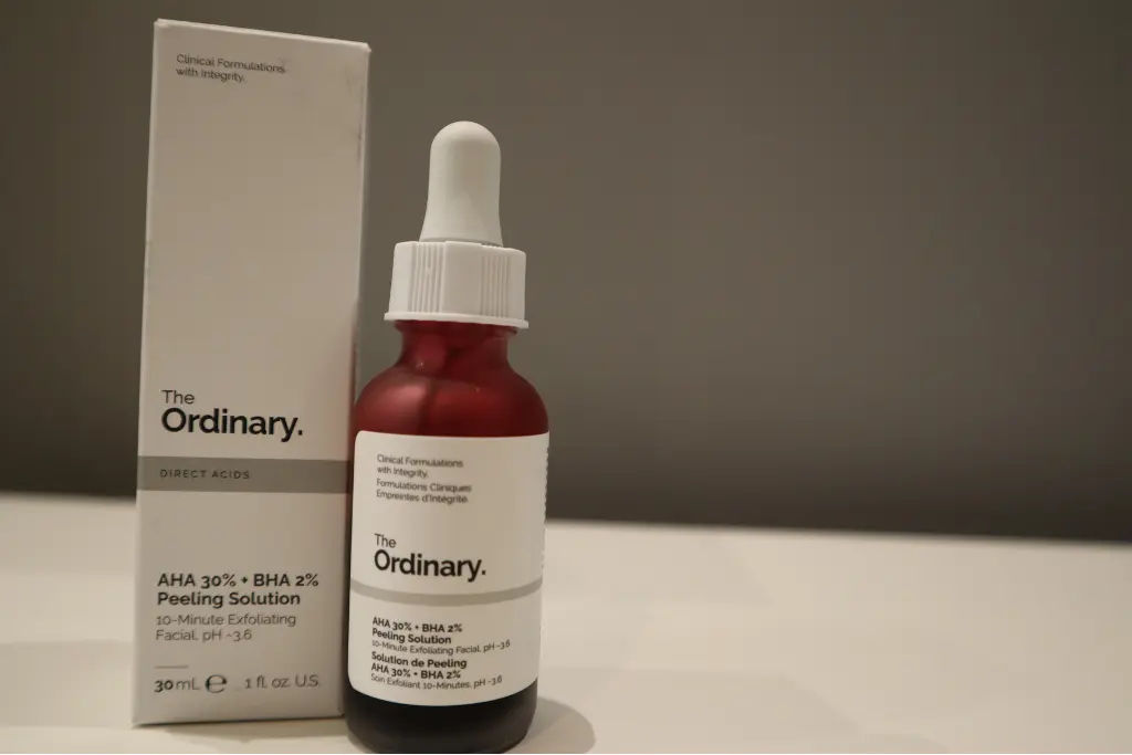 Butelka peelingu The Ordinary stoi obok pudełka na blacie, neutralne szare tło.