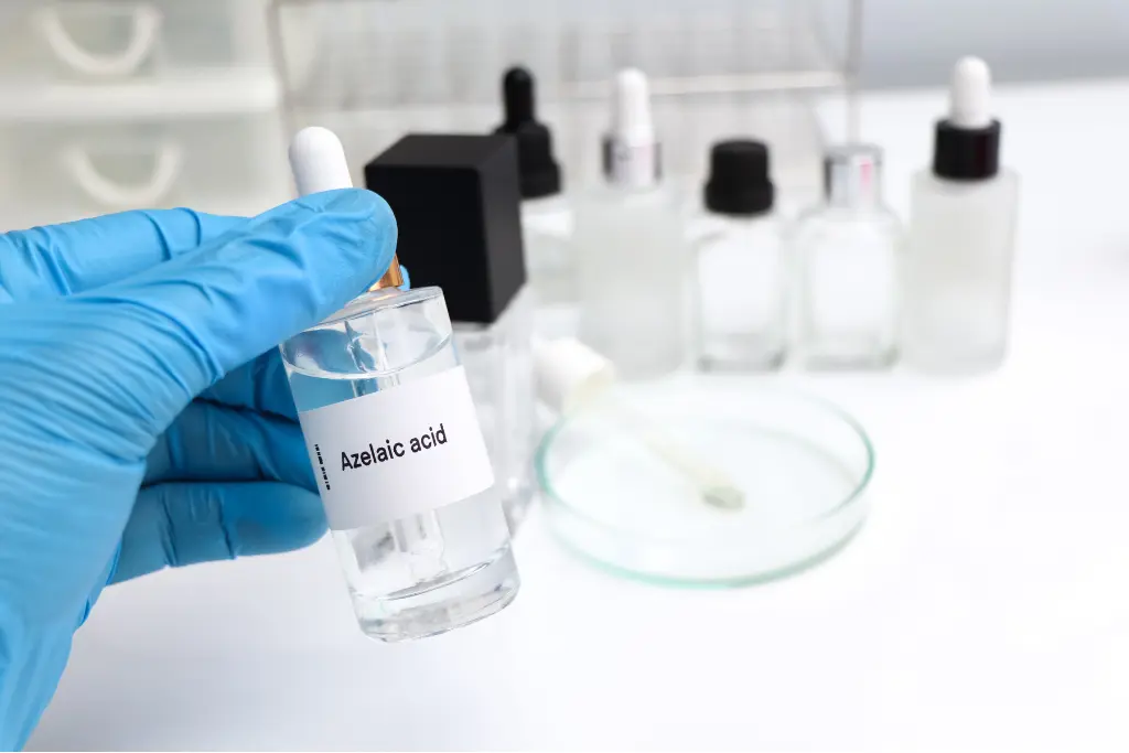 Dłoń w rękawiczce trzyma w laboratorium butelkę z kwasem azelainowym, w tle inne butelki szklane