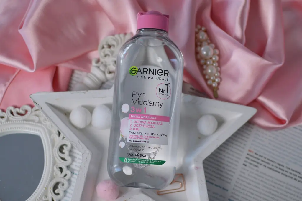 Butelka płynu micelarnego Garnier stoi na białej tacce, otoczona kulkami waty i różowym satynowym tłem.