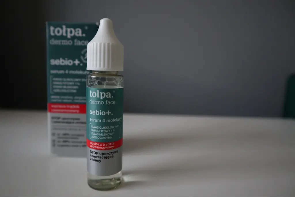 Butelka serum do twarzy stoi na białym blacie, z pudełkiem w tle i szarym tłem.