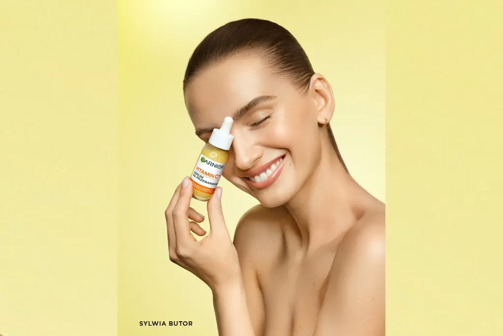 Butelka serum z pipetą trzymana przez uśmiechniętą kobietę na jednolitym żółtym tle.