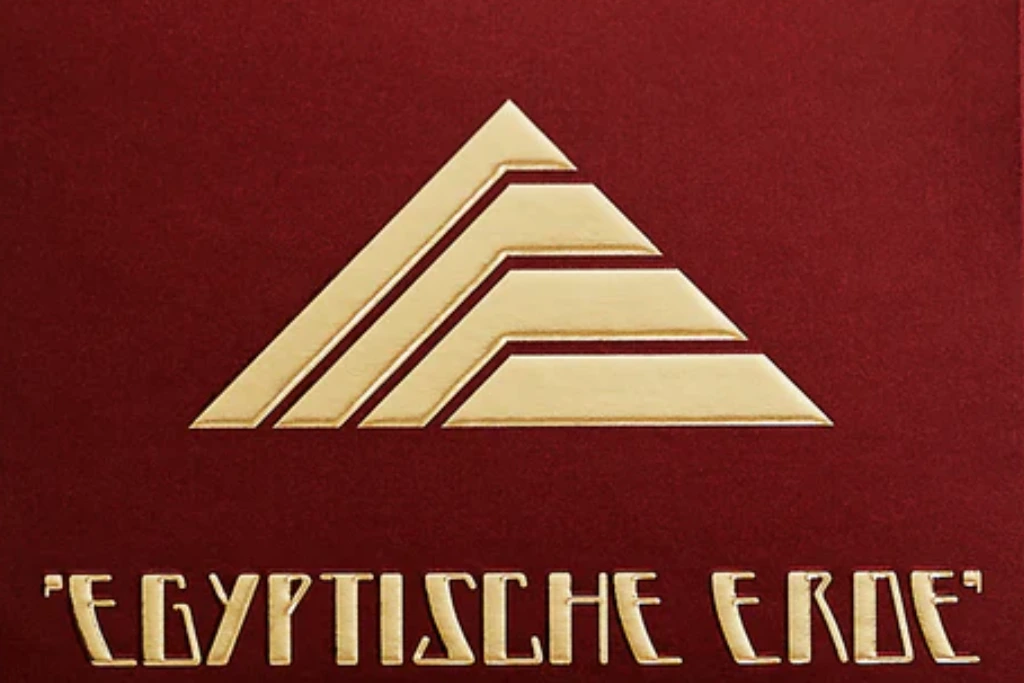 Złote, stylizowane logo piramidy umieszczone nad złotym napisem "EGYPTISCHE ERDE" na bordowym tle.