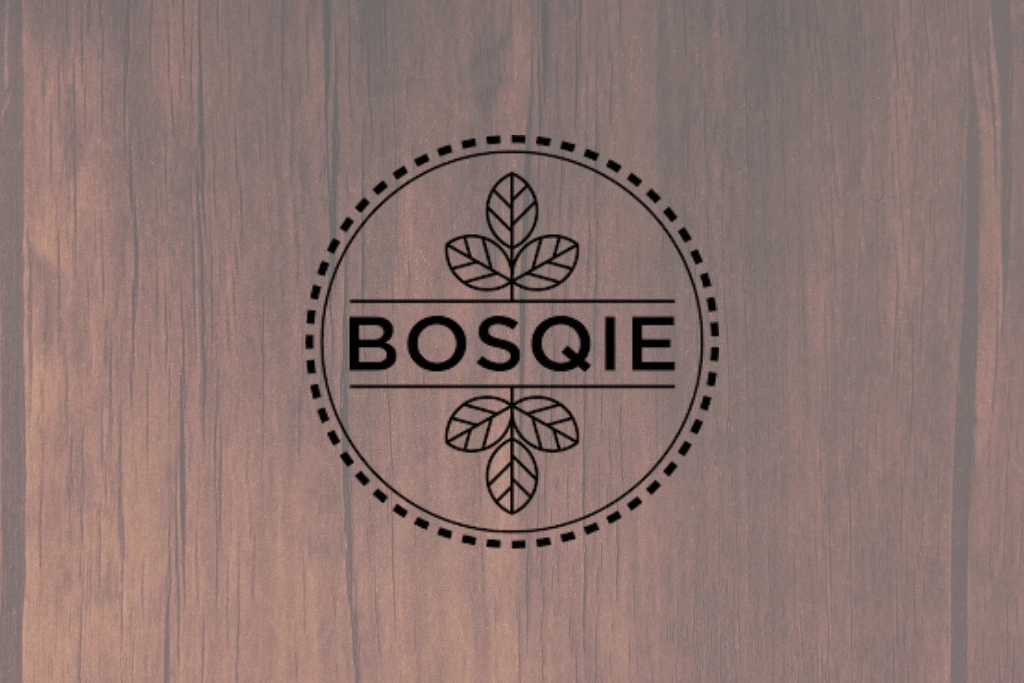 Logo "BOSQIE" nadrukowane na drewnianej tafli, w okrągłej ramce ozdobionej liściowym motywem.