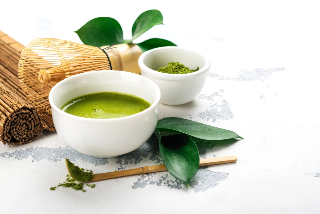 Miseczka z zieloną herbatą matcha, obok mata bambusowa oraz patyczek do rozdrabnianiamatchy