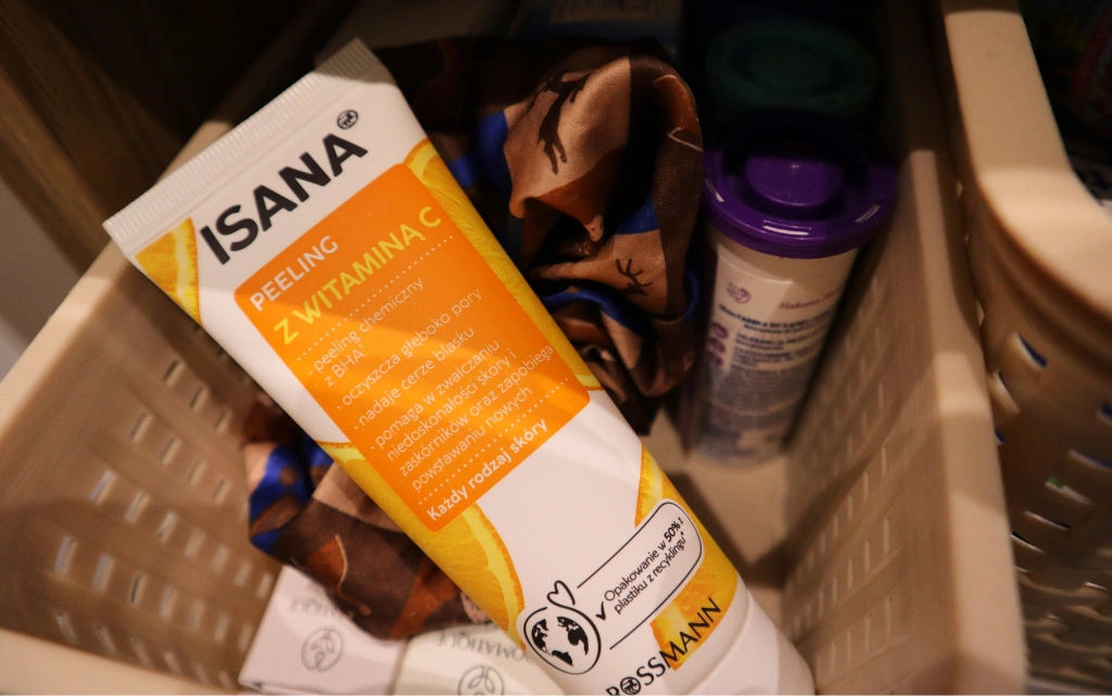 ISANA, peeling do twarzy, z witaminą C w koszyczku w łazience z innymi kosmetykami i gumką do włosów