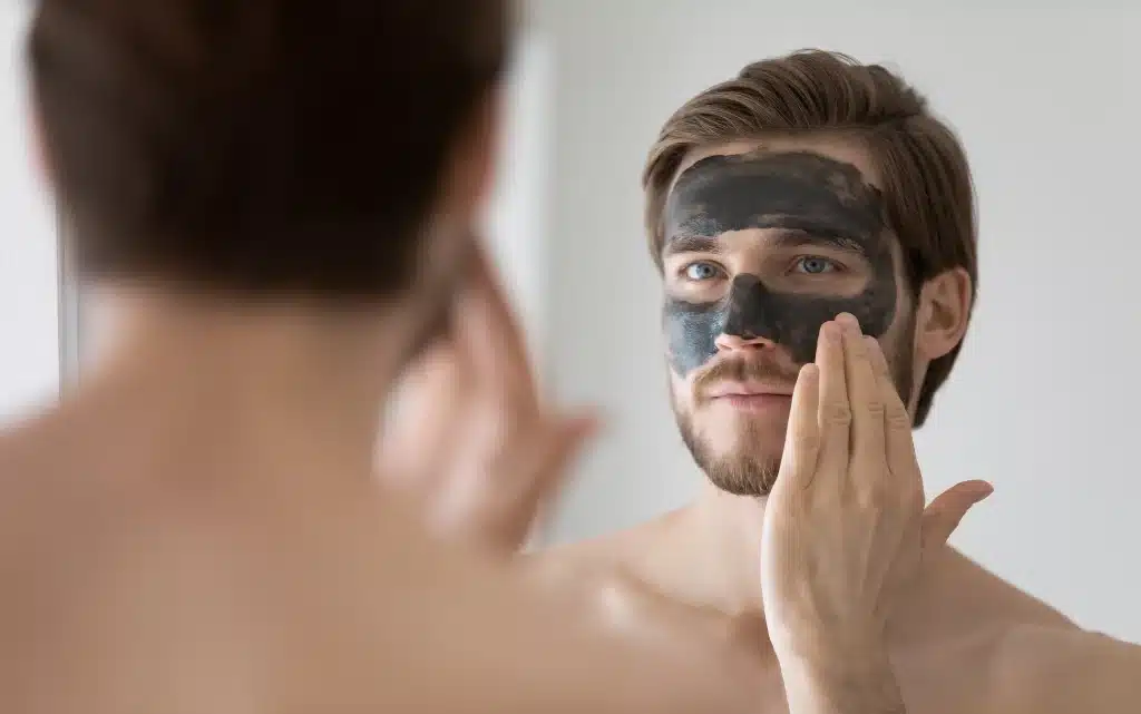 Młody mężczyzna wykonuje peeling twarzy z użyciem kosmetyku z aktywnym węglem
