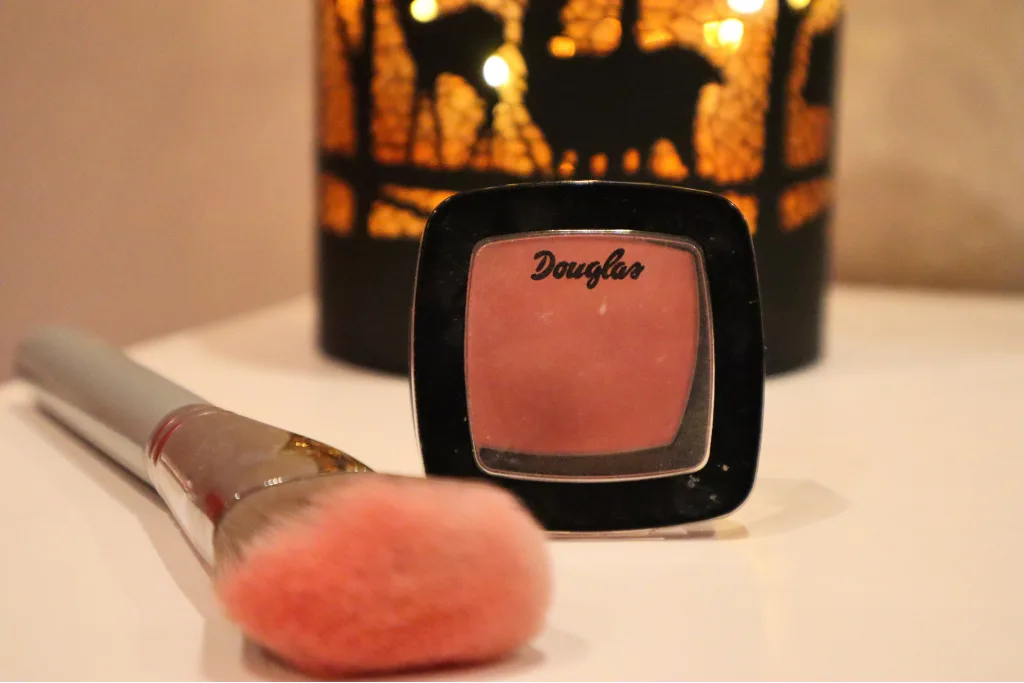 Róż do policzków Douglas Collection Cheek Me Up, obok pędzel do różu, w tle jesienna lampka