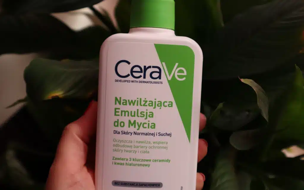 Kobieca dłoń trzyma kosmetyk CeraVe nawilżająca emulsja do mycia dla skóry normalnej i suchej na tle zielonej rośliny 