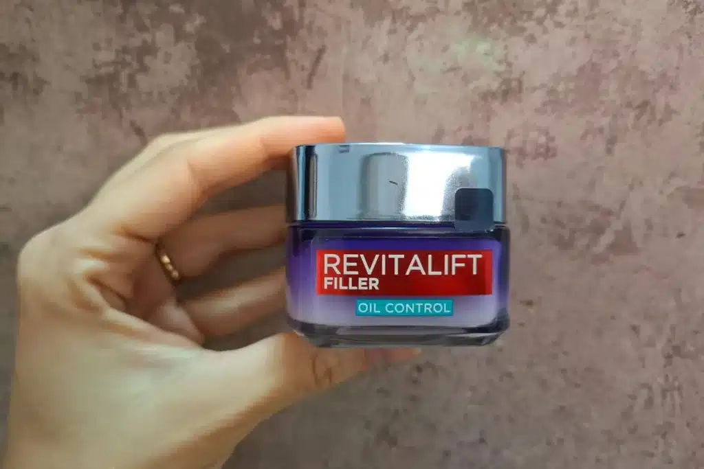 Słoiczek kremu Revitalift Filler Oil Control trzymany dłonią przed różowawą ścianą.