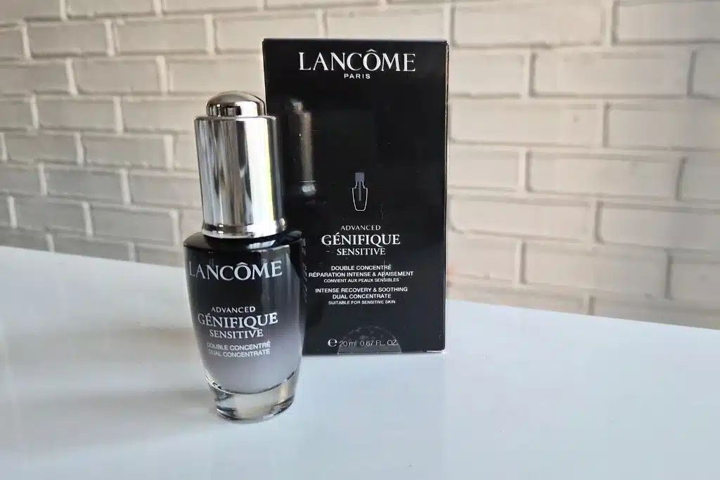 Butelka serum Lancôme stoi obok pudełka na białym blacie przed jasną ceglaną ścianą.