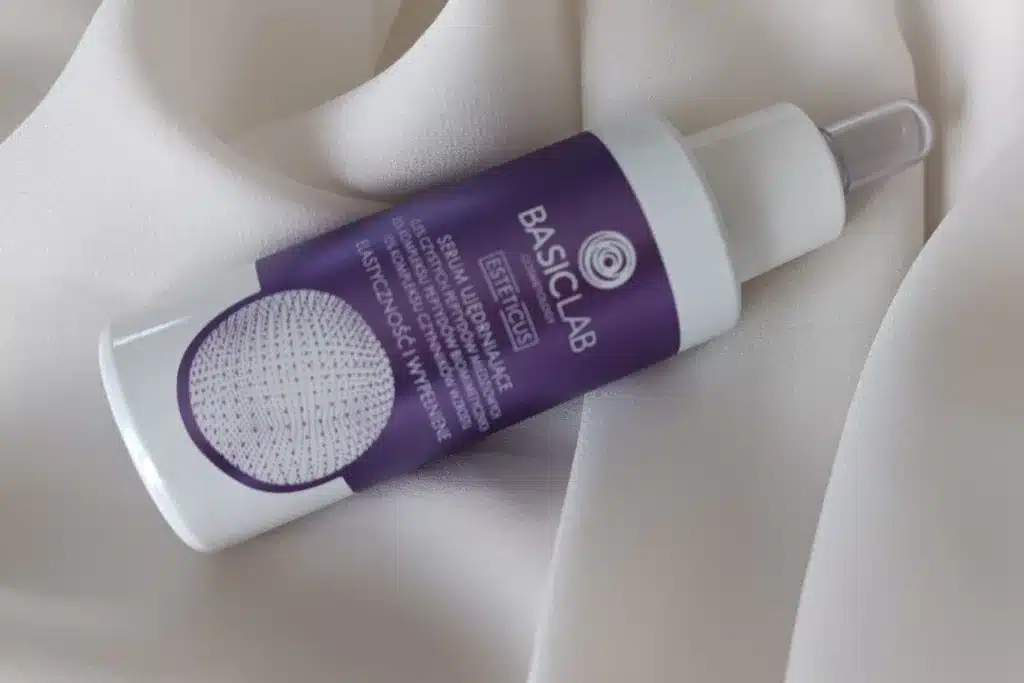 Butelka serum BasicLab z pipetą leży ukośnie na kremowej, marszczonej tkaninie w miękkim świetle.