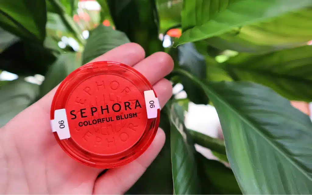W kobiecej dłoni Rozświetlający róż w pudrze Sephora Collection Colorful Blush 06 Tropical Dawn  na tle zielonych liści 