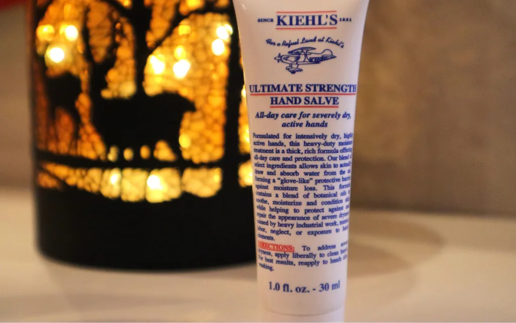 Krem do rąk Kiehl's Ultimate Strenght Hand Salve  na tle świecącej, dekoracyjnej lampki 