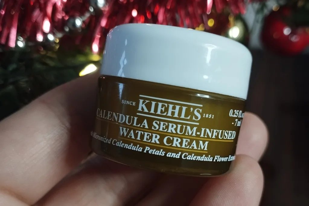 Mały słoiczek kremu Kiehl's Calendula trzymany w dłoni przed rozmytymi świątecznymi ozdobami.